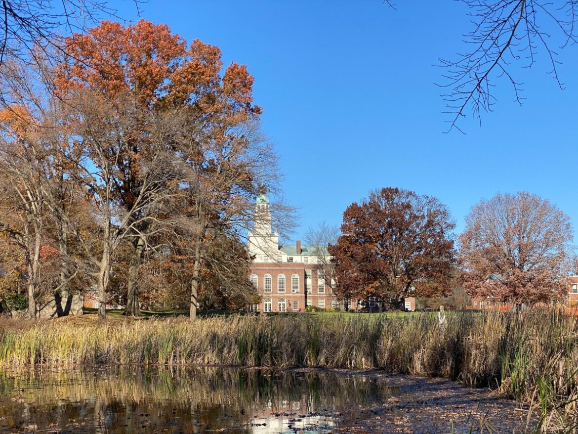A wooded lake, with a stately Georgian brick building visible in the distance.